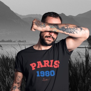 Bărbat tatuat care își acoperă parțial fața cu brațul, purtând un tricou negru cu textul 'PARIS 1980' în roșu și albastru, pe fundal cu peisaj montan alb-negru.