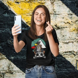 Femeie zâmbitoare purtând un tricou negru cu un design inspirat de cultura hip-hop, ce prezintă un personaj cu o bandană și un radio boombox, pe un fundal colorat, făcând un selfie și gesticulând în fața unui perete colorat.