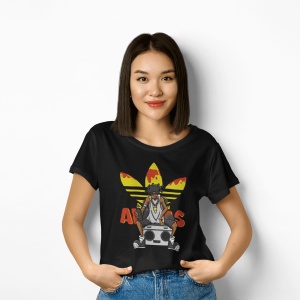 Femeie zâmbitoare purtând un tricou negru cu un design inspirat de cultura hip-hop, ce prezintă un personaj cu o bandană și un radio boombox, pe un fundal grafic colorat, pozând relaxat cu mâinile în buzunari în fața unui perete alb.