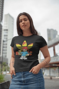 Femeie purtând un tricou negru cu un design grafic colorat ce include logo-ul 'Adidas' stilizat și un personaj desenat(Super Buddy), pozând în aer liber cu clădiri moderne în fundal.