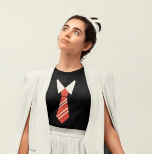 Fată purtând un tricou negru cu un design grafic stilizat de cravată și guler de cămașă, completat cu un sacou alb deschis purtat pe umeri, pe un fundal neutru, uitându-se în sus.