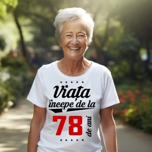 O femeie purtând un tricou alb cu textul  'Viața începe de la 78 de ani'. Fundal din lemn cu diferite nuanțe de maro și verde.