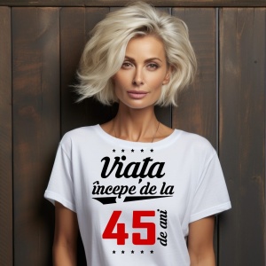 O femeie purtând un tricou alb cu textul  'Viața începe de la 45 de ani'. Fundal din lemn cu diferite nuanțe de maro și verde.