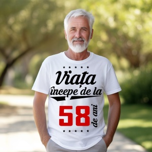 Bărbat în vârstă cu barbă albă, zâmbitor, purtând un tricou alb cu mesajul 'Viața începe de la 58 de ani', stând într-un parc luminat de soare.