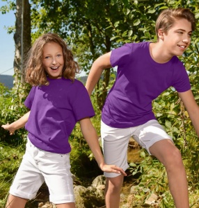 Doi copii, un băiat și o fată, purtând tricouri violet și pantaloni scurți albi, zâmbind și bucurându-se de o zi însorită în natură, înconjurați de copaci și vegetație