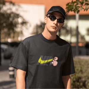 Tânăr cu ochelari de soare și șapcă neagră purtând un tricou negru cu un design Nike stilizat, incluzând un personaj animat lângă logo(Catwoman). Fundal urban ușor neclar, cu elemente de vegetație și clădiri.
