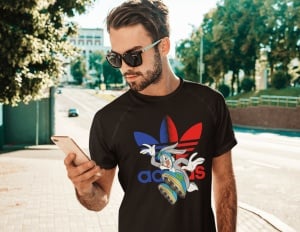 Bărbat purtând un tricou negru cu un design colorat ce include un personaj animat și un logo reinterpretat în culori roșu și albastru, privind telefonul într-un cadru exterior pe o alee însorită.
