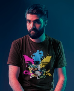 Bărbat cu barbă, purtând un tricou negru cu un design vibrant ce include un personaj animat(Mickey Mouse) și splaturi de culoare roz, albastru și galben, fotografiat într-un cadru cu iluminare artistică în tonuri de albastru și roz.