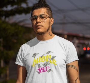 Bărbat cu tatuaje pe față și gât, purtând un tricou alb cu textul 'hustle HARD' și imagini cu bancnote, pe un fundal urban la apus sau în lumina de seară. Poartă ochelari și are o expresie serioasă.