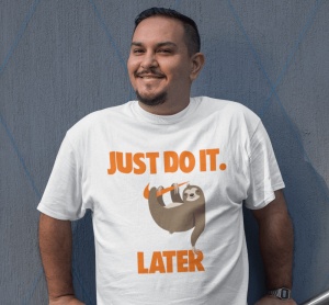 Un bărbat zâmbitor poartă un tricou alb cu un mesaj amuzant în portocaliu care spune 'JUST DO IT. LATER' și o ilustrație a unui leneș agățat de o ramură.