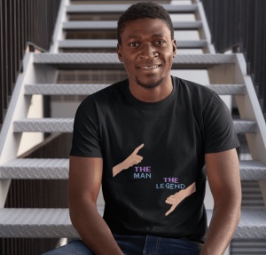 Un bărbat zâmbitor poartă un tricou negru cu un design amuzant ce include două mâini care arată cu degetul, una în sus cu textul 'The Man' și una în jos cu textul 'The Legend'. Bărbatul stă pe o scară metalică, fundalul fiind un cadru industrial.