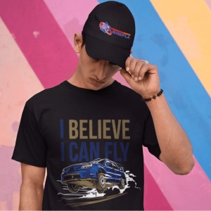 Bărbat purtând un tricou negru cu un design grafic al unei mașini sport albastre în aer și textul 'I BELIEVE I CAN FLY', alături de o șapcă albă inscripționată cu logo-ul 'Imbracaminte Simplă', pe un fundal colorat cu dungi diagonale.