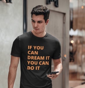 Bărbat purtând un tricou negru cu mesajul motivațional 'IF YOU CAN DREAM IT YOU CAN DO IT' scris cu litere portocalii, ținând un telefon în mână și mergând printr-un spațiu modern de interior.