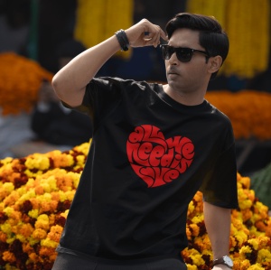Bărbat purtând ochelari de soare și un tricou negru cu un design roșu în formă de inimă, conținând textul 'All we need is love', stând în fața unui fundal de flori portocalii și galbene.
