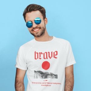 Un bărbat zâmbitor poartă ochelari de soare rotunzi și un tricou alb cu inscripția 'brave' în roșu, însoțită de o imagine grafică cu un soare și un peisaj montan stilizat. Fundalul este un perete albastru vibrant, adăugând contrast și evidențiind designul