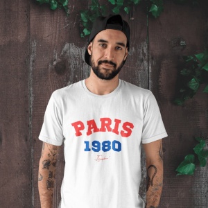 Bărbat cu barbă purtând o șapcă neagră pe spate și un tricou alb cu textul 'PARIS 1980' în roșu și albastru, pe fundal de lemn decorat cu frunze verzi.
