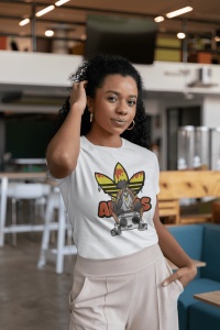 Femeie purtând un tricou alb cu un design inspirat de cultura hip-hop, ce prezintă un personaj cu o bandană și un radio boombox, pe un fundal grafic colorat, pozând într-un spațiu modern de interior, cu mâna ridicată la păr.