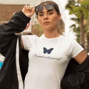 Fată serioasă purtând un tricou alb cu un fluture albastru și textul 'Be gentle with yourself', ridicându-și ochelarii de soare pe cap. Pozează afară într-o zonă urbană, purtând o jachetă deschisă.