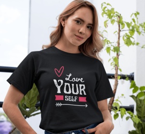 Fată purtând un tricou negru cu un design grafic ce conține textul 'Love Yourself', alături de un desen cu inimă și săgeată, într-un cadru luminos cu plante în fundal.