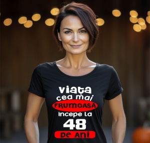 Femeie zâmbitoare purtând un tricou negru cu mesajul în română: 'Viața cea mai frumoasă începe la 48 de ani', pe un fundal estompat cu lumini.