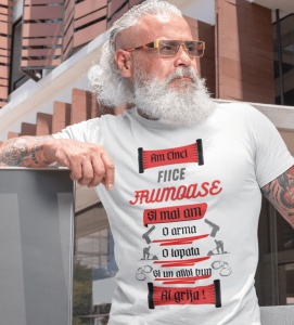 Bărbat cu barbă albă și păr lung, purtând un tricou alb cu mesajul 'Am cinci fiice frumoase și mai am o armă, o lopată și un alibi bun. Ai grijă!', alături de ilustrații cu arme, o lopată și cătușe. Bărbatul pozează în aer liber lângă o clădire modernă.
