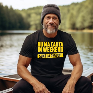 Un bărbat matur, zâmbitor, purtând un tricou negru cu mesajul "Nu mă căuta în weekend, sunt la pescuit" scris cu galben. Stă pe o barcă, într-un peisaj liniștit cu lac și pădure în fundal, purtând o căciulă de lână.
