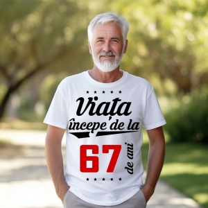 Bărbat în vârstă cu barbă albă, zâmbitor, purtând un tricou alb cu mesajul 'Viața începe de la 67 de ani', stând într-un parc luminat de soare.
