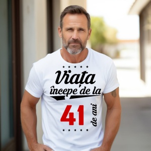 Bărbat matur cu barbă scurtă, purtând un tricou alb cu mesajul 'Viața începe de la 41 de ani', stând în exterior pe o alee însorită.