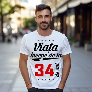 Bărbat zâmbitor purtând un tricou alb cu mesajul 'Viața începe de la 34 de ani', stând pe o stradă urbană însorită.