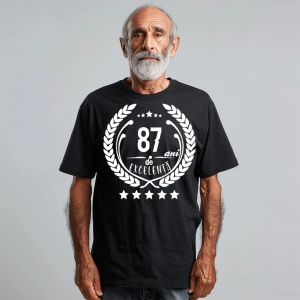 Un bărbat zâmbitor purtând un tricou negru cu textul ''87 de ani de excelență'', înconjurat de o coroană de lauri.
