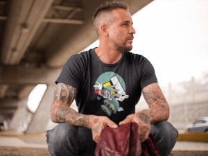 Bărbat tatuat purtând un tricou negru cu un design grafic colorat, stând pe vine sub o structură de pod modern, cu privirea îndreptată în lateral și ținând o geacă în mâini.
