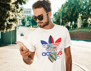Bărbat purtând un tricou alb cu un design colorat ce include un personaj animat și un logo reinterpretat în culori roșu și albastru, privind telefonul într-un cadru exterior pe o alee însorită.