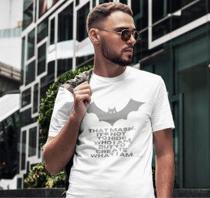 Bărbat purtând ochelari de soare și un tricou alb cu un design inspirat de Batman, având o siluetă de liliac și textul: 'THAT MASK. IT'S NOT TO HIDE WHO I AM, BUT TO CREATE WHAT I AM.' Fundalul este urban, cu o clădire modernă din sticlă și metal în spate