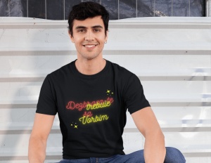 Un bărbat zâmbitor, cu păr negru și ondulat, stă așezat și poartă un tricou negru cu un mesaj colorat. Textul de pe tricou, scris în nuanțe de roșu și galben neon, spune 'Dezbracă-te să vorbim,' fiind accentuat cu mici stele decorative galbene. Fundalul e