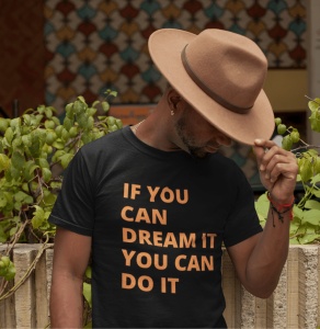 Bărbat purtând un tricou negru cu mesajul motivațional 'IF YOU CAN DREAM IT YOU CAN DO IT' scris cu litere portocalii și o pălărie maro, înclinând capul în jos. Fundalul include o plantă și un perete decorativ cu modele geometrice colorate.