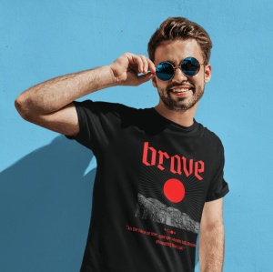 Un bărbat zâmbitor poartă ochelari de soare rotunzi și un tricou negru cu inscripția 'brave' în roșu, însoțită de o imagine grafică stilizată cu un soare și un peisaj montan. Fundalul este un perete albastru vibrant, iar bărbatul ridică ușor ochelarii cu 