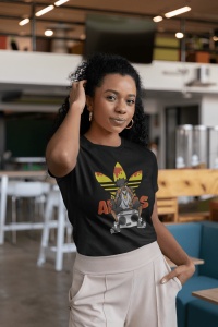 Femeie purtând un tricou negru cu un design inspirat de cultura hip-hop, ce prezintă un personaj cu o bandană și un radio boombox, pe un fundal grafic colorat, pozând într-un spațiu modern de interior, cu mâna ridicată la păr.