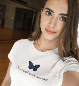 Fată zâmbitoare purtând un tricou alb cu un fluture albastru și mesajul 'Be gentle with yourself'. Fotografia este un selfie făcut în interior, cu fundal estompat, ce sugerează o cameră spațioasă.