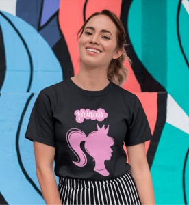 Tânără purtând un tricou negru cu un design roz care înfățișează silueta unei prințese cu o coroană și cuvântul 'princess' deasupra. Ea zâmbește în fața unui perete viu colorat, decorat cu forme abstracte în nuanțe de albastru, verde și portocaliu.
