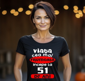 Femeie zâmbitoare purtând un tricou negru cu mesajul în română: 'Viața cea mai frumoasă începe la 51 de ani', pe un fundal estompat cu lumini.