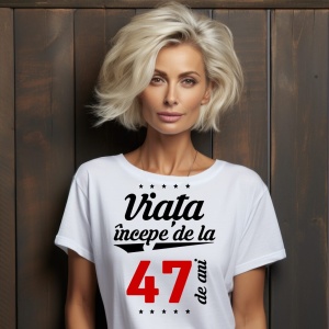 O femeie purtând un tricou alb cu textul  'Viața începe de la 47 de ani'. Fundal din lemn cu diferite nuanțe de maro și verde.
