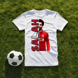 Tricou alb personalizat cu imaginea fotbalistului Salah și text vertical roșu cu numele 'Salah', așezat pe iarbă lângă o minge de fotbal.