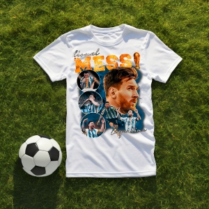 Tricou alb cu un design dedicat lui Lionel Messi, incluzând imagini ale fotbalistului și textul 'Messi Argentina', așezat pe iarbă alături de o minge de fotbal.
