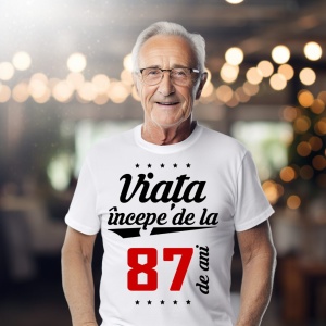 Bărbat în vârstă purtând ochelari și un tricou alb cu mesajul 'Viața începe de la 87 de ani', într-un cadru iluminat cald.